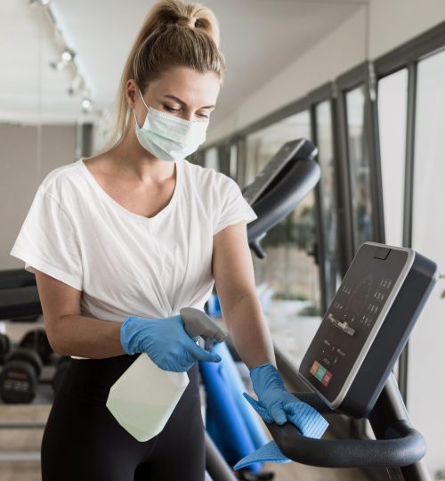 mulher-com-luvas-limpando-equipamento-de-ginastica-enquanto-usa-mascara-medica